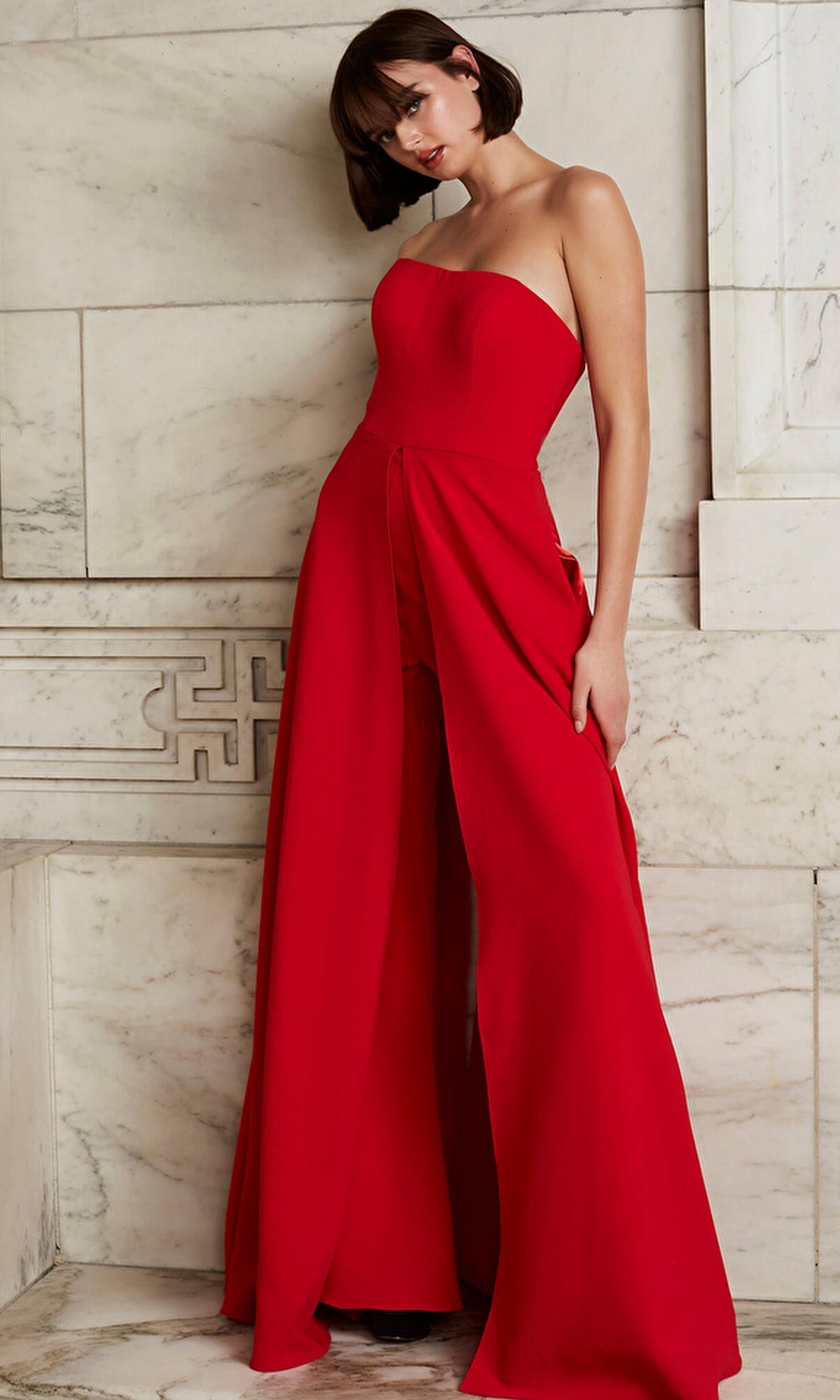 A person stands against a marble wall wearing the Jovani Strapless Wide-Leg Formal Jumpsuit 03529—a bold red, strapless jumpsuit with wide legs and a chic front overlay.